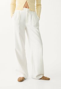 Witte wijde broek met plooien, gemaakt van een gladde stof. Gecombineerd met beige instap schoenen met metallic accenten.