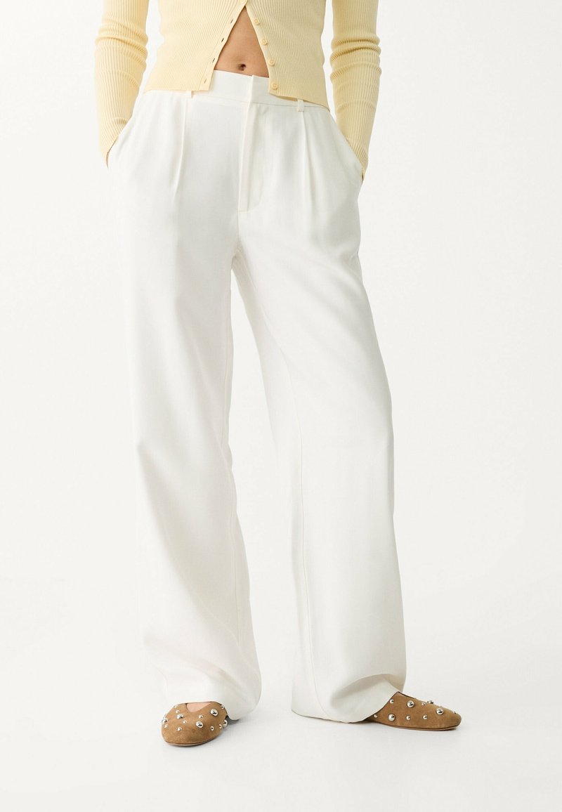 Witte wijde broek met plooien, gemaakt van een gladde stof. Gecombineerd met beige instap schoenen met metallic accenten.