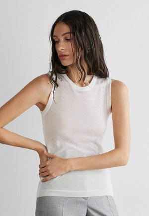 Femme aux longs cheveux bruns portant un haut blanc sans manches et un pantalon gris clair, posant avec les mains près de la taille sur un fond blanc.