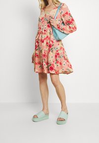 Žena v růžových květovaných šatech s dlouhými rukávy, držící světle modrou kabelku přes rameno a nosící světle zelené platformové sandály na klínku.