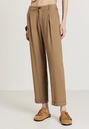 Tan pleated trousers with a relaxed fit, rolled cuffs, and a button closure. Paired with brown fringed moccasin-style shoes.