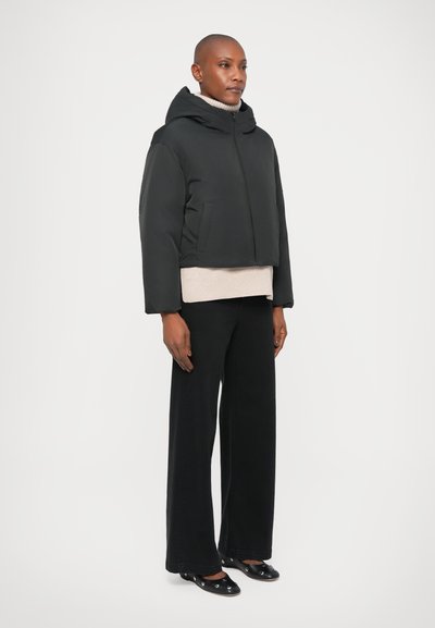 Chaqueta acolchada negra recortada con capucha, con cierre frontal de cremallera y bolsillos laterales. Combinada con un suéter beis y pantalones de pierna ancha negros.