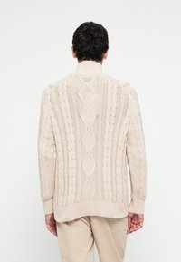 Hombre con suéter de punto trenzado beige y pantalones de color claro, de pie de espaldas contra un fondo blanco liso.