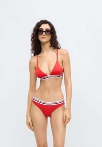 Κόκκινο μπικίνι με τριγωνικό τοπ και πλαϊνές ρίγες σε μαύρο, άσπρο και κόκκινο. Το μοντέλο φοράει σκουρόχρωμα γυαλιά ηλίου και έχει κατσαρά μαλλιά.