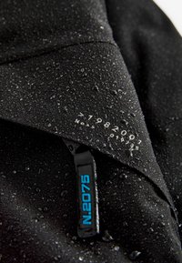 Indumento impermeabile nero con gocce sulla superficie, dotato di un colletto cucito e un'etichetta con la scritta "N.2076" e numeri di serie.