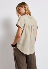 Blusa corta beige chiaro a maniche corte con colletto e dettagli plissettati sul retro, caratterizzata da un orlo curvo. Indossata con pantaloni marroni larghi.
