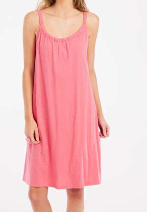 Kobieta ubrana w luźną, bezrękawnikową różową sukienkę na cienkich ramiączkach, stojąca na białym tle.
