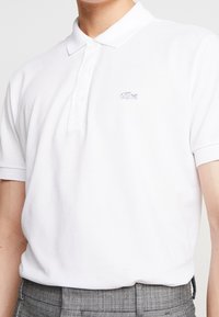 Polo blanc en coton, doté d'une patte à trois boutons et d'un petit logo brodé sur le côté gauche de la poitrine. Manches courtes.