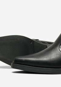 Svart läderankelhatt med spetsig tå, texturerad yta och gummisula. Har elastiska sidopaneler och en subtil logotyp på sidan.