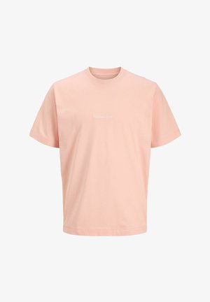 Tenåringsgutt står og ser mot høyre, iført lys rosa T-skjorte, svarte shorts med hvit tekst, kremfargede sokker og hvite joggesko med svarte striper.