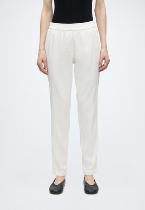 Femme portant un pantalon blanc à taille élastique et des chaussures plates noires, debout devant un fond uni gris clair.