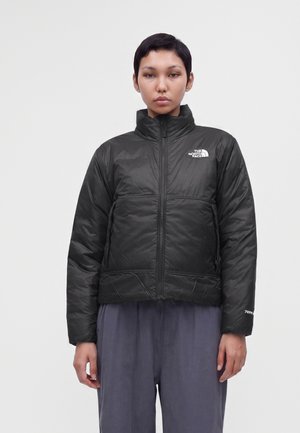Άτομο που φοράει μαύρο μονωμένο μπουφάν The North Face με ψηλό γιακά, στέκεται μπροστά σε απλό φωτεινό φόντο.