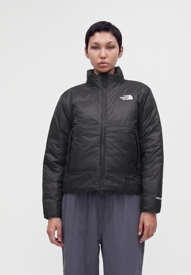 Άτομο που φοράει μαύρο μονωμένο μπουφάν The North Face με ψηλό γιακά, στέκεται μπροστά σε απλό φωτεινό φόντο.