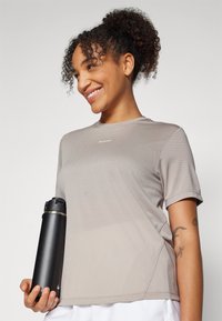 Camiseta atlética de color beige claro con mangas cortas y tejido texturizado. Una botella de agua negra sostenida contra el cuerpo añade contraste.