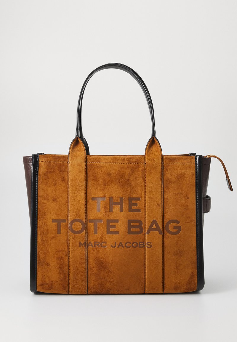 Brązowa torebka tote z zamszu i skóry z czarnymi uchwytami, pionowymi przeszyciami i napisem "THE TOTE BAG MARC JACOBS" nadrukowanym z przodu.