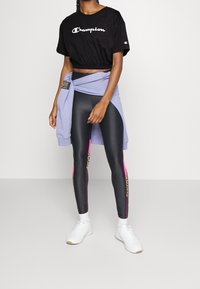 Černé zkrácené tričko s logem "Champion", levandulová mikina uvázaná v pase, černé legíny s růžovými a leopardími akcenty, bílé tenisky.