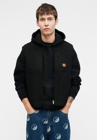 Μαύρο γιλέκο με φερμουάρ και τσέπη καγκουρό, φοριέται πάνω από μια μαύρη κουκούλα, συνδυασμένο με μπλε τζιν με σχέδιο του γιν-γιανγκ.