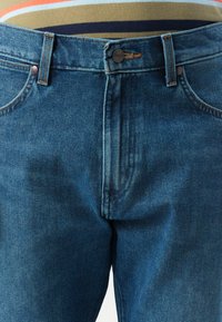 Jean en denim dans un délavé bleu foncé avec une texture légèrement usée. Présente une fermeture à bouton en métal et des coutures orange.