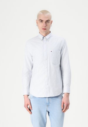 Young man with short blonde hair wears light blue striped button-up shirt and light blue jeans, standing against a plain white background.