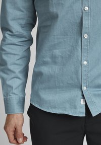 Light blue button-up shirt made of cotton, with a textured finish, long sleeves, and a curved hem, featuring white buttons and a small tag.