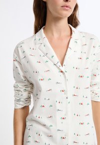 Witte blouse met een kleurrijk patroon van kleine huisjes en bomen. Het materiaal lijkt zacht, met een losse pasvorm en opgerolde mouwen.