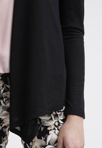 Schwarzer Langarm-Cardigan mit weicher Textur, Drapé-Design und abgerundetem Saum. Kombiniert mit einem pinken Oberteil und floral gemusterten Hosen.