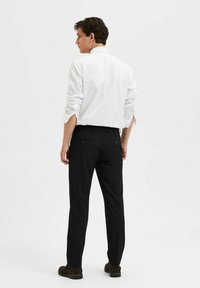 Witte overhemd met knopen, gedragen met zwarte broeken, voorzien van achterzakken. De outfit heeft een strakke pasvorm met opgerolde mouwen en donkere schoenen.
