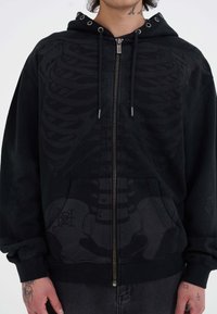 Sudadera negra con cremallera y estampado gráfico de esqueleto, tejido texturizado, capucha con cordón y ojales de metal, y bolsillos frontales.