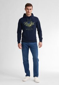 Felpa navy con cappuccio e coulisse, tasca frontale e stampa grafica in verde chiaro. Abbinata a jeans blu e sneakers bianche. Vestibilità casual.