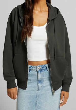 Femme portant un sweat à capuche gris foncé zippé sur un débardeur blanc court et une jupe en denim bleu clair.