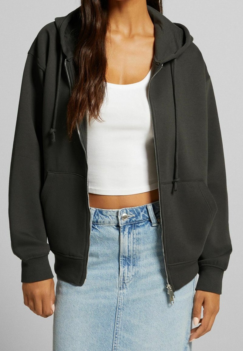 Femme portant un sweat à capuche gris foncé zippé sur un débardeur blanc court et une jupe en denim bleu clair.