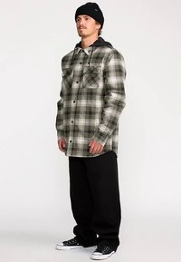 Karierte Flanellhemd in Grün, Schwarz und Weiß mit zwei Brusttaschen, Knopfleiste und schwarzem Hoodie darunter. Getragen mit schwarzer Hose.