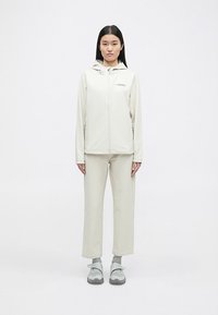 Light beige soft-shell jacket and pants set. Jacket features a hood and zippers; relaxed fit with subtle logo; smooth texture. Lightweight footwear.