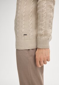Beige Kabelstrickpullover mit geripptem Saum und Bündchen; verfügt über ein dezentes schwarzes Markenzeichen an der Seite. Kombiniert mit hellbraunen Hosen.