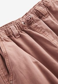 Pudrosiai rožiniai šortai su surinkta elastinga juosta ir raišteliu, plokščiais šoninėmis kišenėmis, pagaminti iš lengvos, švelnios medžiagos.