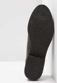 Semelle de chaussure noire avec motif de bande de roulement texturé, inclinée contre un mur blanc d’angle, affichant la pointure 37 sur la semelle.