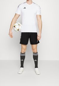 Valkoinen lyhythihainen urheilupaita, jossa on mustia raitoja, yhdistettynä mustiin shortseihin ja mustiin sukkiin, joissa on valkoisia raitoja. Pelaaja pitää kädessään moniväristä jalkapalloa.