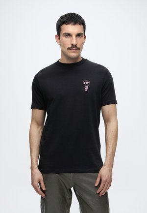 Homme aux cheveux courts et foncés avec moustache portant un t-shirt noir avec un petit motif rose et un pantalon gris, debout sur fond blanc.