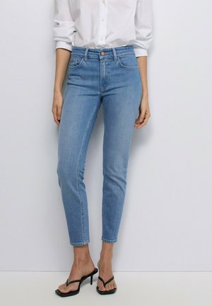 Mujer con vaqueros pitillo de talle alto de color azul claro, una camisa blanca abotonada con las mangas remangadas y sandalias negras de tacón.