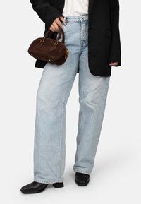 Persona vistiendo jeans anchos de pierna en azul claro, botas negras de punta afilada, blazer negro y sosteniendo un pequeño bolso marrón.