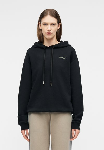 Γυναίκα με καστανά ίσια μαλλιά που φοράει μαύρο φούτερ Off-White και μπεζ παντελόνι, στέκεται μπροστά σε απλό ανοιχτό γκρι φόντο.