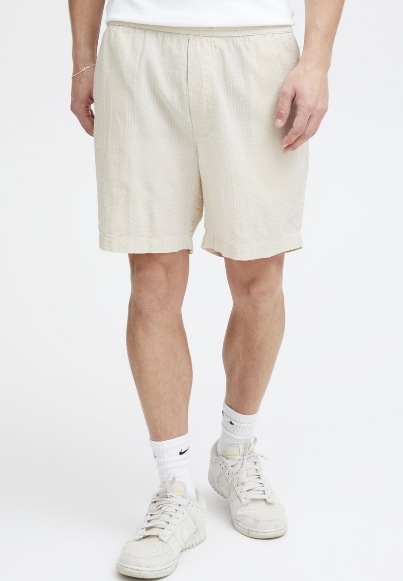 Beige shorts med strukturmønster, elastisk midjebånd og sidelommer, brukt med hvite sneakers og ankellange sokker.