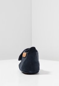 Ciemnoniebieskie kapcie o miękkiej zamszowej strukturze, okrągłym nosku i małym brązowym skórzanym logo blisko pięty. Prosty, stonowany design.