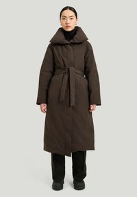 Brun lang jakke med bred krave, bælte i taljen og lange ærmer, parret med en sort turtleneck og sorte bukser, der har en glat tekstur.
