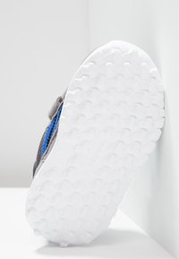 Harmaa urheilukenkä sinisin yksityiskohdin. Kenkässä on valkoinen, teksturoitu kumipohja, jossa on kuusikulmainen kuvio. Sivukuva korostaa kengän muotoilua.