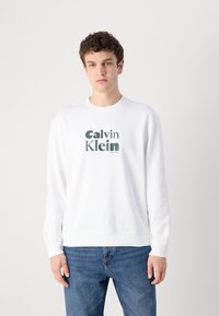 Biely crewneck sveter vyrobený z bavlny, s zeleným vytlačeným logom v strede. Dlhé rukávy a rebrované manžety a lem.