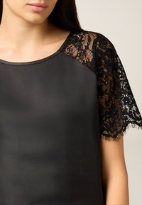 Czarna bluzka z gładkim materiałem na ciele i krótkimi rękawami z czarnej koronki, ozdobiona misternymi wzorami i frędzlami na krawędziach.