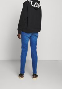Zwarte hoodie met witte tekst op de rug, gecombineerd met blauwe denim jeans en sandalen met beige zool. Gladde, casual uitstraling.