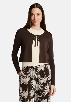 Frau trägt eine dunkelbraune, kurz geschnittene Strickjacke über einem cremefarbenen Top und braune Hose mit weißem Palmenmuster, steht vor einem weißen einfarbigen Hintergrund.