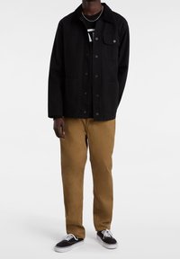 Mężczyzna w czarnej zapinanej kurtce, czarnej koszulce z grafiką, beżowych spodniach i czarnych niskich trampkach z białymi podeszwami i sznurowadłami, stojący.